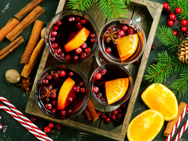 4 vague christmas cocktails on a wood tray, surrounded by evergreen branches, candy canes, berries, and cinnamon bark 